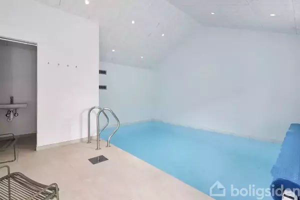 Indendørs swimmingpool med lyseblå vand fylder rummet. Der er en stige til at træde ned i poolen. Flisebelagt gulv og hvidmalede vægge omgiver poolen.