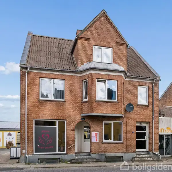 En murstensbygning med skarpt tag har store vinduer i stueetagen. En hvid varevogn holder parkeret til venstre. Skiltning udenfor viser en stiliseret rose og teksten "in zone."