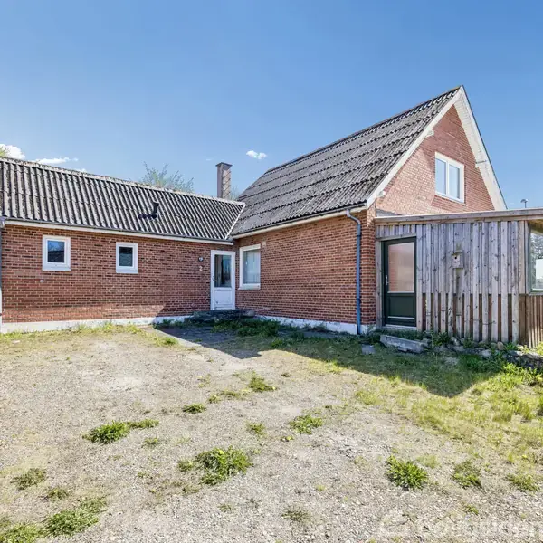 Et rødt murstenshus står stille på en åben, grusbelagt grund. Ved siden af er en lille tilbygning med træpaneler. Omkring ligger der græs og et hegn.