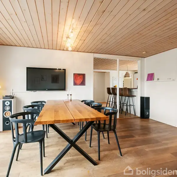 Et træbord står midt i stuen omgivet af sorte stole. Et fladskærms-tv hænger på væggen. Stuen har trægulv, træloft og adgang til en balkon gennem store glasdøre.