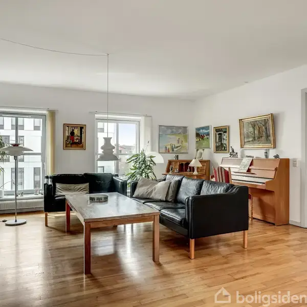 Stue med sort lædersofa og træstuebord. Et klaver står i hjørnet. Rummet har trægulve, kunst på væggene og store vinduer, der giver naturligt lys.