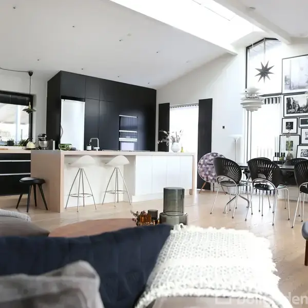 Stue med moderne køkkenø, sorte og hvide stole omkring spisebord, samt vægdekorationer. Sofa og tæppe i forgrunden, store vinduer og højt til loftet i lyst rum.