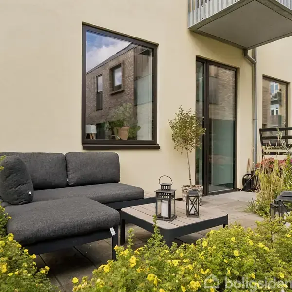 Sofa står på terrasse omgivet af grønne planter. Ved siden af er der et lille bord med to lanterner. Baggrunden viser en moderne bygning med store vinduer.