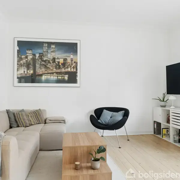 En beige sofa står i en stue ved siden af et rundt, sort lænestol. Et stort billede af en by i natten hænger på væggen. En fladskærm er monteret på væggen over et hvid skab med planter og kasser.