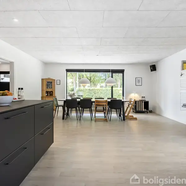 Køkken-alrum med sort køkkenø fyldt med appelsiner, lys stue med lang spisebord, sorte stole, stor vindue. Hvide vægge med hylder fyldt med bøger og billeder.
