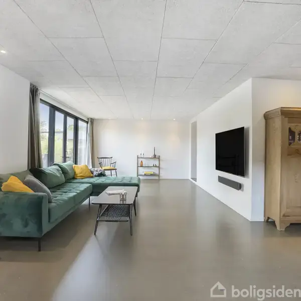 Grøn sofa med gule puder står ved et stort vindue. Et sofabord foran, en væghængt tv-skærm og en træskab i hjørnet. Lys, rummelig stue med minimalistisk indretning.