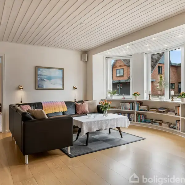 En stue med sort sofa og sofabord dækket med dug. Store vinduer viser huse udenfor, og reoler fyldt med bøger fylder det buede hjørne. Lampen lyser vægmaleri.