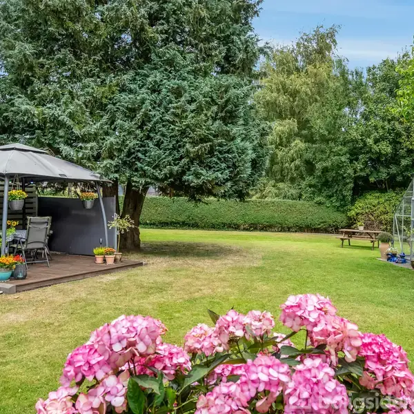 Pavillon står i en have omgivet af grønne træer og buske. En nærliggende drivhus står til højre, mens pink hortensia blomstrer i forgrunden.