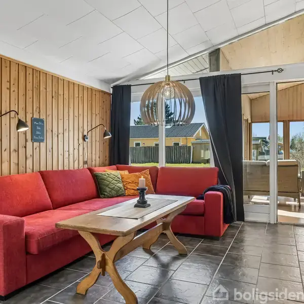 Rød sofa står i en stue med træpanelvægge og mørkt flisegulv. Et træbord foran sofaen. Store vinduer med sorte gardiner giver udsigt til solfyldt terrasse.