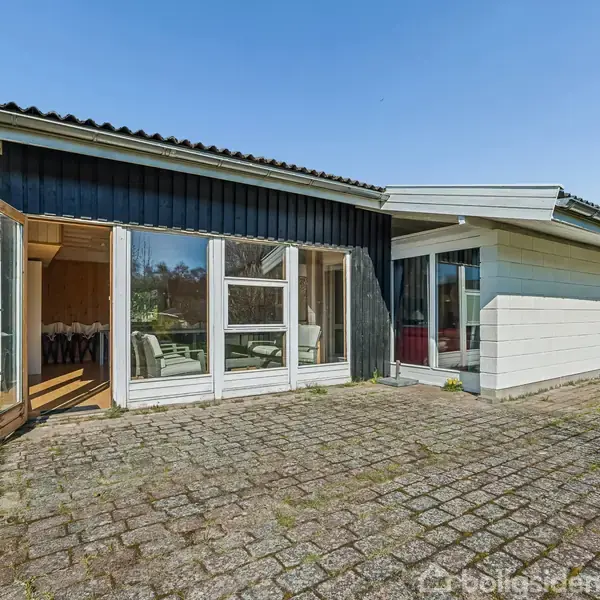 Hus med store vinduer og åbne døre, inviterer lys ind i en rummelig stue. Beliggende på en flisebelagt terrasse omgivet af græsplæne under en klar blå himmel.
