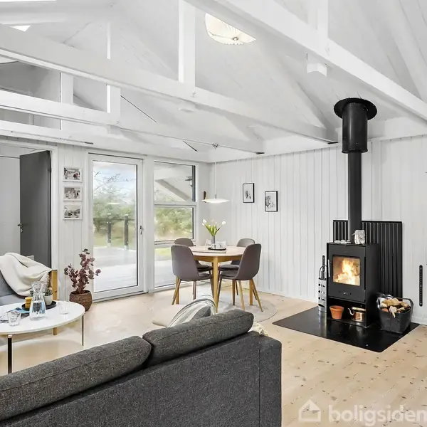 En brændeovn opvarmer en stue. En sofa og et rundt sofabord står til venstre. Et spisebord med to stole er ved siden af et vindue, der viser udsigt til natur.