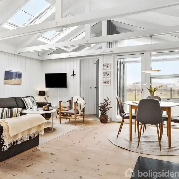 Sofa står i en lys stue med trægulve og hvide vægge. Et spisebord med fire stole er ved vinduet. Loftet har synlige bjælker og ovenlysvinduer. Pejs til højre.
