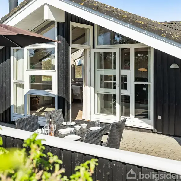 Sort sommerhus med store vinduer og skråt tag. Terrasse med udendørs spisebord, parasol, og stole. Omgivet af natur med træer i baggrunden.