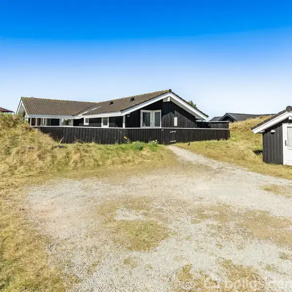 Et sort træhus med hvide vinduesrammer står på en grusvej omgivet af klitter og græsarealer i et åbent landskab under en klar blå himmel. En mindre bygning ses til højre.