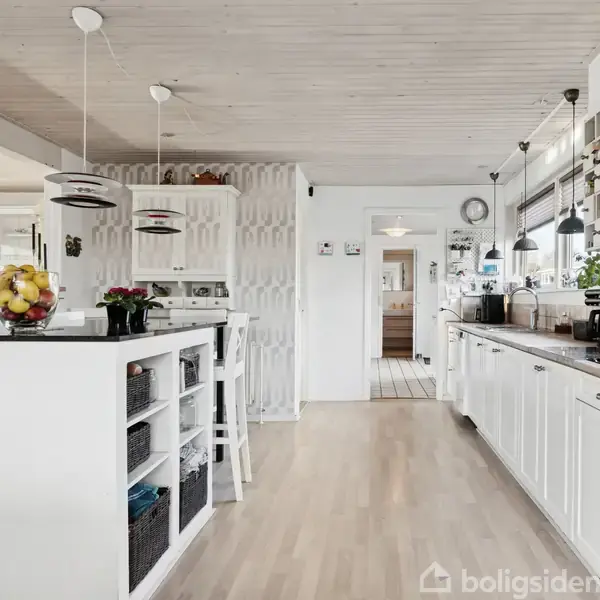 Et moderne køkken med hvide skabe og en lang bordplade fyldt med køkkenudstyr. Frugtskål og barstole står ved en halvmur. Lyse trægulve og hængende lamper skaber en lys atmosfære.