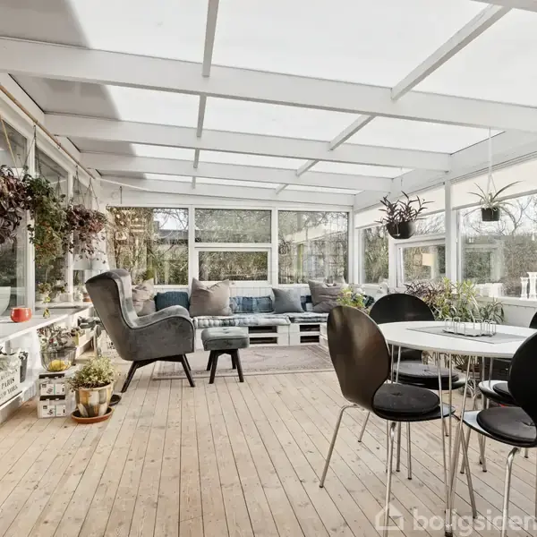 En udestue med lyse trægulve har et bord med fire sorte stole som central objekt. Potteplanter hænger fra loftet og står på hylderne langs vinduerne i venstre side.