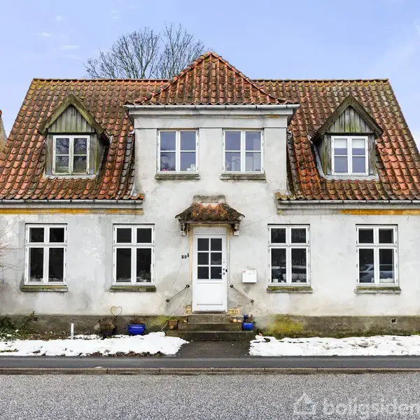 Huset har to etager med fire vinduer foran, et centralt udhæng og en rød tegltag. Det står ved en gade, om vinteren, med sne på fortovet. Nummeret "87" ses ved døren.