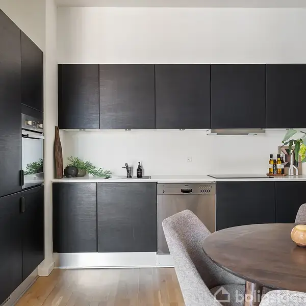 Et moderne køkken med sorte skabe og stålprodukter. Køkkenet ligger op ad en hvid væg med træplankegulv. En rund spisebord dekoreret med blomster står i forgrunden.