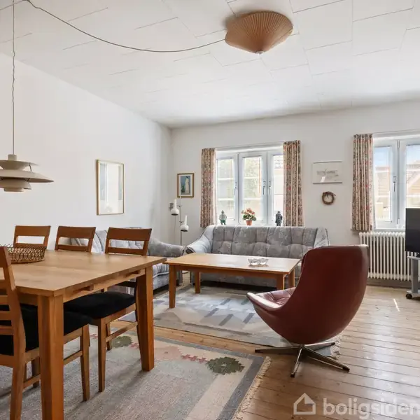 Et spisebord med stole står i en lys stue. Rummet har en sofa ved vinduerne, en rød lænestol, et tv på et stativ og træmøbler på trægulv.
