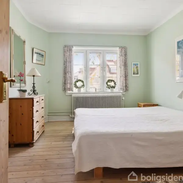 Dør åbner ind til et soveværelse med en seng dækket af hvidt sengetøj. Til venstre står en trækommode med en lampe. Værelset har lysegrønt interiør og et vindue med gardiner.