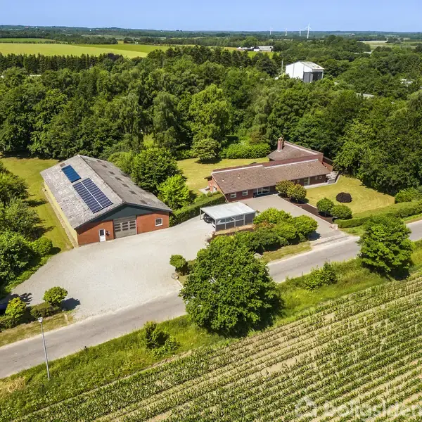 Luftfoto af en landejendom med en stor lade med solpaneler, en rød murstenshus og en gårdsplads omgivet af frodig skov og marker i det solrige landskab.