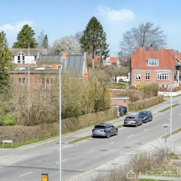 En stille vej med parkerede biler, omkranset af ældre, charmerende huse med røde tegltage, omgivet af grønne haver og træer, under en klar blå himmel.