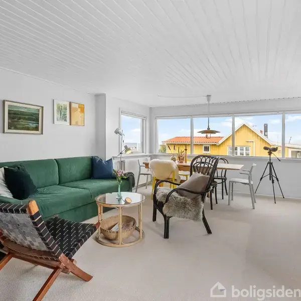 En stue med grøn sofa og pejs til venstre. To stole står ved et lille sofabord. Et spisebord med stole står ved de store vinduer med udsigt til et gult hus.