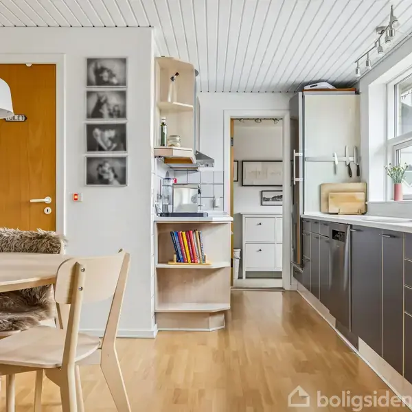 Et køkken med træbord og stole til venstre, en hvid lampe hænger over bordet. Til højre er der mørke køkkenskabe og en opvaskemaskine. Vinduet giver udsigt til træer.