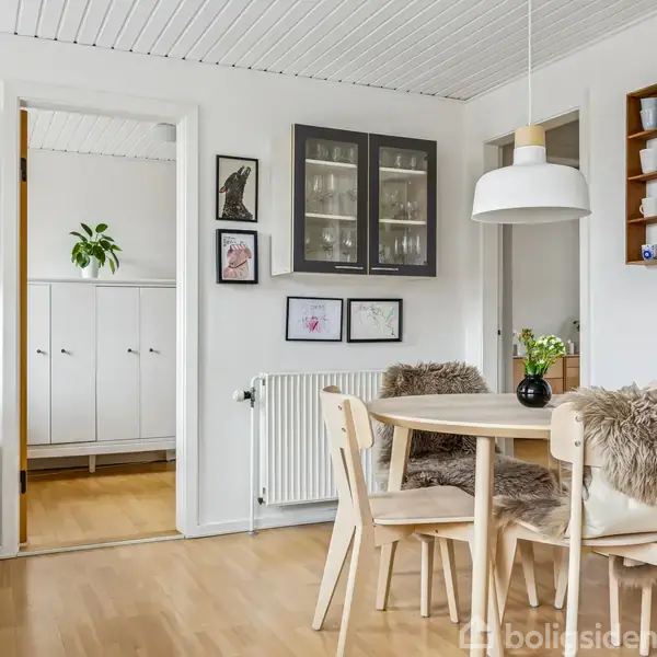 Rundt spisebord med fire træstole dækket af fåreskind, vase med blomster, hænger under en hvid pendellampe. Væghylder med porcelæn og kunst pryder væggene i et lyst køkkenmiljø.