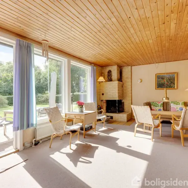 Stue med træloft og store vinduer, der lader sollys ind. Møbleret med polstrede stole og sofabord, pejs til højre. Yderligere udsigt til terrasse og grønt udenfor.