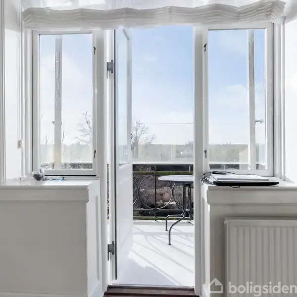 Åben dør fører ud til en altan med sort metalbord og stol. Hvide vægge og radiator omgiver scenen med et billede på væggen.