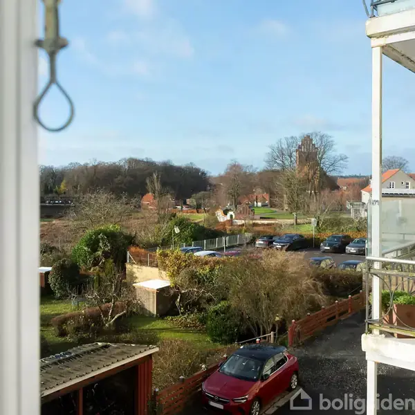 En rød bil holder parkeret foran en bygning med altaner. I baggrunden ses en række huse og parkerede biler omgivet af træer på en klar dag.