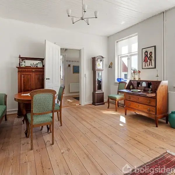 Stue med træbord og grønne stole ved en sofagruppe. En antik kommode, en standerur og vægpynt i baggrunden. Lyse trægulve og vinduer skaber et lyst rum.