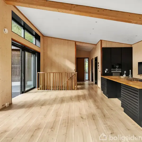 Et lyst køkken-alrum med trægulv og træloft, store vinduer til venstre, en køkkenbordsø med sort base til højre, står i et åbent moderne rum med skovudsigt.