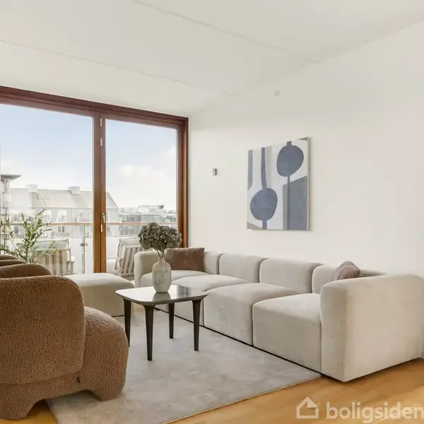 En stue med en lysegrå modulsofa og brune lænestole. Sofaen står på et tæppe foran et lavt bord. Store vinduer giver udsigt til en balkon og bygninger udenfor.