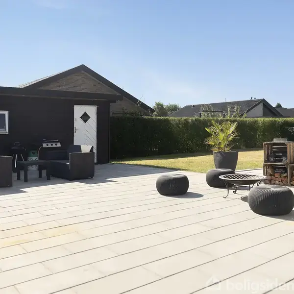 Terrassemøbler står på en rummelig flisebelagt gårdhave. Flere sorte puffer omkranser et lavt bord. I baggrunden er der en græsplæne med omgivende hæk og et sort skur.