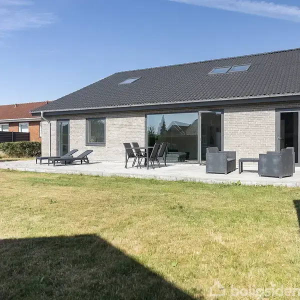 En moderne, enetages murstensvilla med mørkt tag ses fra baghaven. Villaen har en terrasse med loungemøbler og liggestole. Græsplæne omgiver huset i et forstadsområde.