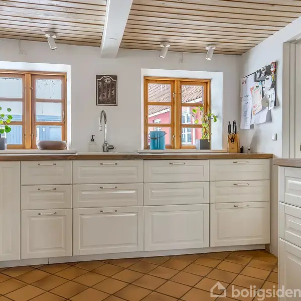 Køkkenbord med vask og hvide skabe; planter og køkkenredskaber på bordet. To vinduer med udsigt til en rød bygning. Lysebrune fliser og trætæppe på loftet.