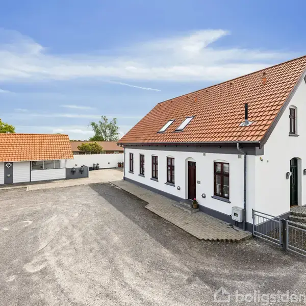 Hvidt hus med rødt tegltag står stille i et åbent gårdspladsområde omgivet af lave bygninger og spredte træer under en klar blå himmel.