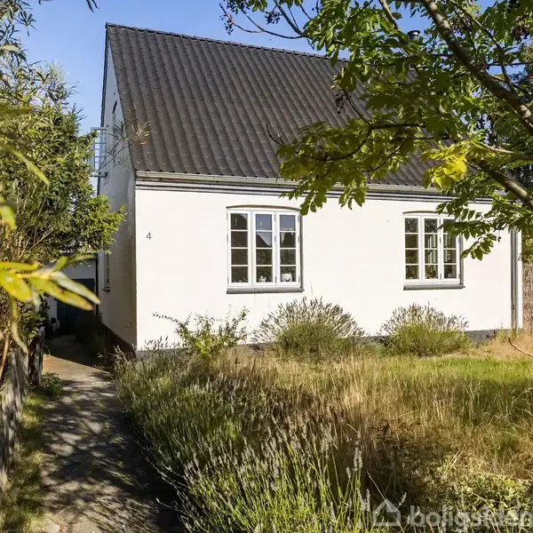 Hus står stille, omgivet af en have med græs og buske. Huset har hvid facade og mørkt tag med to vinduer foran. Tallet "4" ses på væggen.