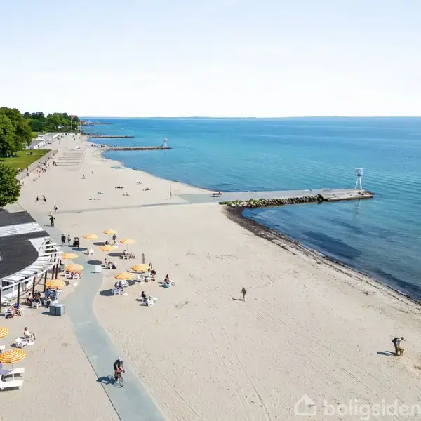 Strand med gyldent sand, folk solbader og går, omgivet af klart havvand og grønne træer. Spredte parasoller og en promenade langs stranden.