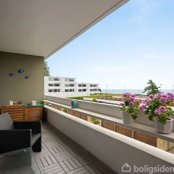 Altan med mørke møbler og blomster i potter står stille. Den har udsigt over et moderne boligkompleks og havet i baggrunden under en blå himmel.