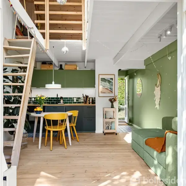 Trætrappe fører til hems i stue med grøn sofa og spisebord. Køkkenet er bagved med grønne skabslåger. Væggene prydes af planter og kunstværker i lyse omgivelser.