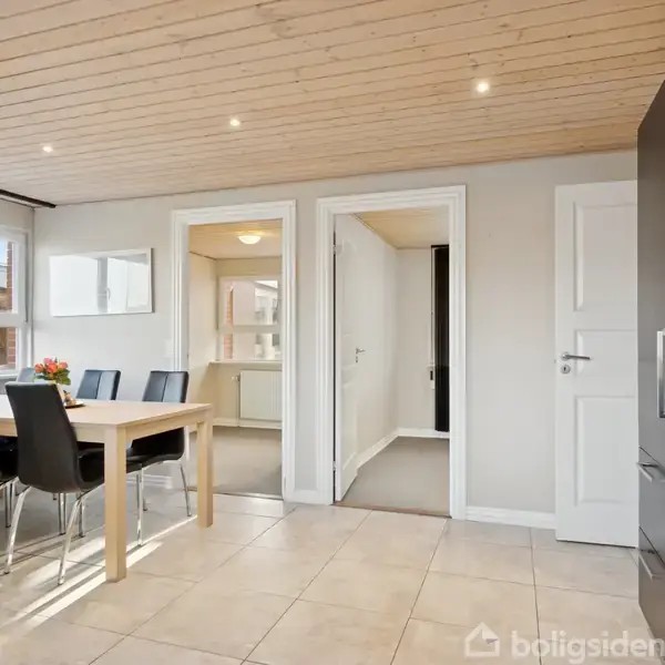 Et lyst og moderne køkken med fliser, sort indbygget ovn og mikrobølgeovn. Der er et spisebord med sorte stole og vindue i baggrunden, alt omgivet af hvide vægge og døre.