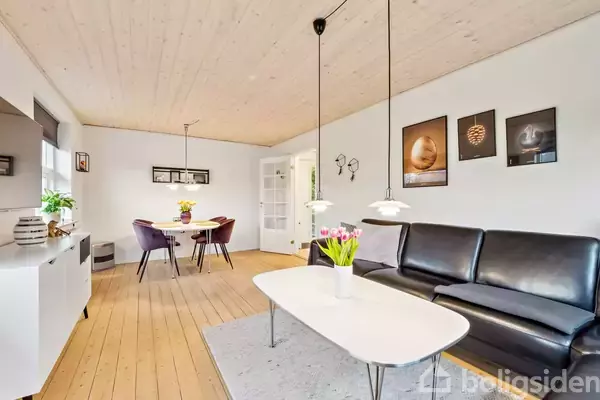 En stue med lædersofa og sofabord med blomster. Spisebord med lilla stole står i baggrunden. Rummets vægge har kunstværker, og træloftet er lyst. Gulvet er træ.