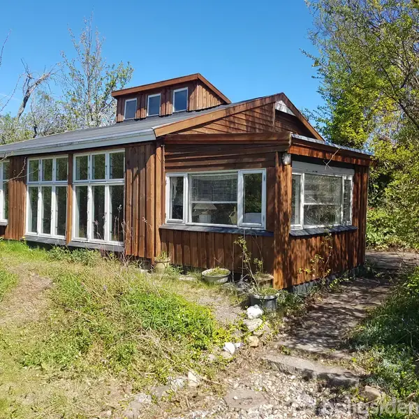 Et træhus med store vinduer står i en frodig have. Solen skinner, og der er grønne træer og buske omkring. Huset har et opbygget loftsvindue.