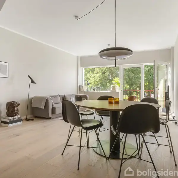 En rund spisebord med sorte stole står i en stue. Ved siden af er en sofa og et gulvlampe. Store vinduer giver udsigt til grønne træer udenfor.