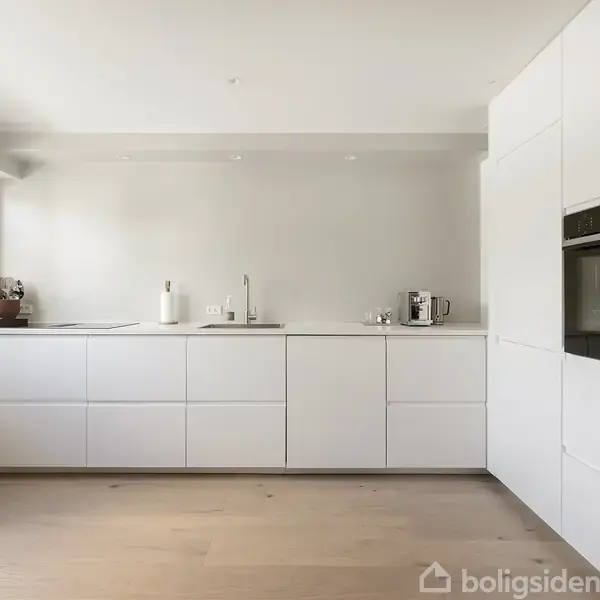 Et lyst, minimalistisk køkken med hvide skabe og indbyggede apparater. På bordet står en blender og kaffemaskine. Store vinduer til venstre slipper dagslys ind. Gulvet er i lyst træ.