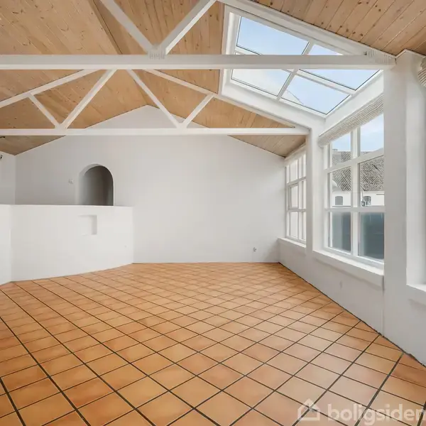 Rummeligt værelse med terrakottafliser på gulvet, stort skråt loft med synlige bjælker. Store vinduer og glastag slipper masser af lys ind. Væggene er hvide, og der ses en rundbuet indgang.