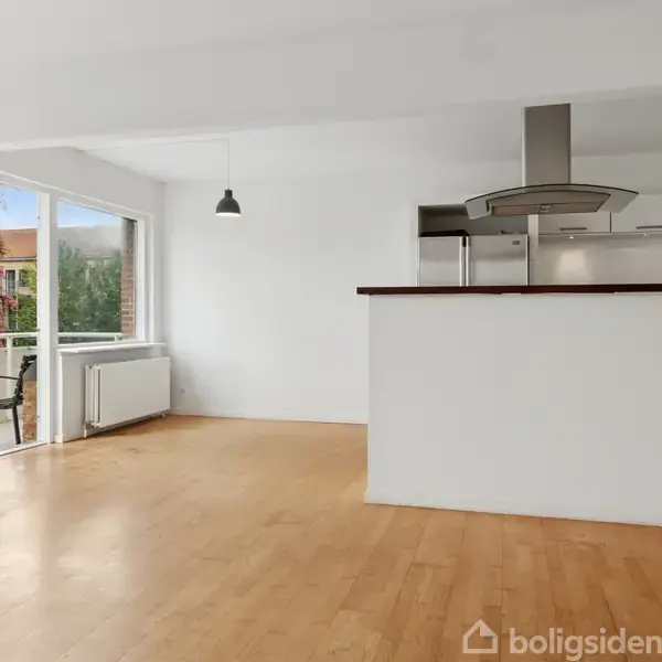 Et lyst stueområde med trægulve og en åben forbindelse til et moderne køkken. Store vinduer og en dør fører ud til en balkon med udsigt til grønne omgivelser.